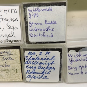 May include: A collection of small, clear plastic boxes, each containing a specimen and a handwritten label. The labels include information such as the mineral name, location, and collector's name. The boxes are arranged in a white container.
