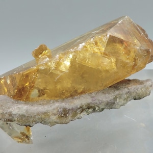 Puede incluir: Un espécimen mineral cristalino de color amarillo dorado y translúcido que presenta una punta afilada y alargada. El cristal está unido a una base de matriz rugosa, de color claro y texturizada, con tonos tierra y bordes irregulares.