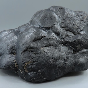 May include: A dark gray, irregularly shaped rock with a rough, textured surface. The rock has a slightly metallic sheen and appears to be a single, solid piece. The overall form is organic and natural, with rounded edges and a dense appearance.