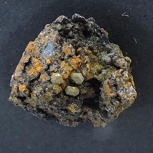 Op de afbeelding: Een ruw, onregelmatig mineraalexemplaar met een donkere, poreuze en metaalachtig ogende matrix met vlekken van geoxideerde oranjebruine roest. In de holtes zijn verschillende kleine, bleek olijfgroene, zeshoekige kristallen ingebed.