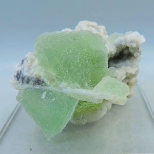 Puede incluir: Un grupo de cristales de fluorita verde pálido con una superficie texturizada, sobre un fondo de formaciones minerales blancas y grises. La fluorita tiene una cualidad translúcida, y la composición general es natural y orgánica.