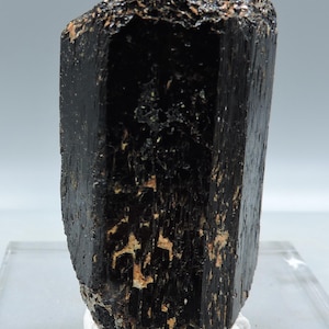 May include: A black tourmaline crystal with a rough, textured top and a polished, striated body. The crystal is displayed on a white base, showcasing its dark color and natural form. The mineral has a cylindrical shape.