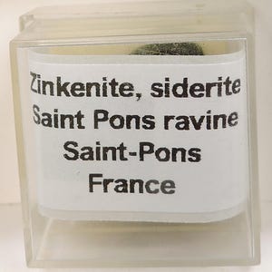 May include: A small, clear plastic display case containing a mineral specimen. The label reads "Zinkenite, siderite Saint Pons ravine Saint-Pons France". The mineral appears dark green and textured.