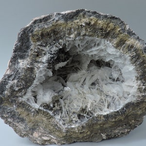 May include: A geode with a cavity lined with clear, white, needle-like crystals. The geode is surrounded by a dark grey, green, and brown rock.