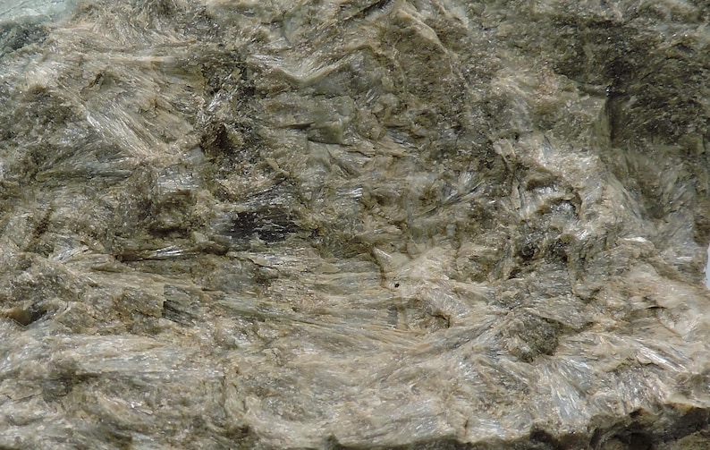 May include: Close-up of a textured rock formation with a mix of gray and light green hues. The surface has a layered appearance with crystalline structures and rough, uneven textures. The overall look is natural and earthy.
