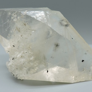 May include: A clear quartz crystal with a faceted, geometric form. The quartz features internal inclusions and a rough, textured base. The overall colour is translucent white with dark specks. The crystal is set against a neutral background.
