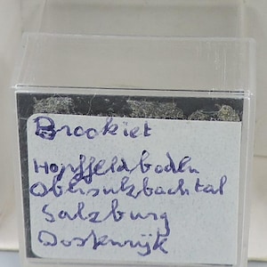 Puede incluir: Una pequeña caja de plástico transparente que contiene un espécimen. Una etiqueta blanca dentro de la caja tiene texto manuscrito en tinta azul que dice "Brookiet", seguido de varias líneas de texto.