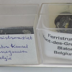 May include: Two small, clear plastic display cases containing mineral specimens. One case holds a dark, textured rock with handwritten text: "Ferristrunziet, Blaton Kanaal, Henegouwen, Belgie." The other case has a mineral labeled "Ferristrunzite Mont-des-Groseille, Blaton, Belgium."