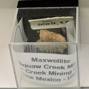 May include: A small, light-coloured mineral specimen, possibly Maxwellite, encased in a clear plastic display box. The box has a black top and a white label with text indicating the mineral's origin in New Mexico, USA.