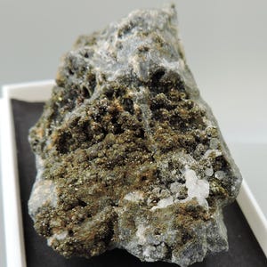 May include: A rough, gray rock with patches of sparkling, golden-brown mineral deposits. Small, clear crystal clusters are visible on the surface. The rock is displayed in a white tray, showcasing its natural texture and color variations.