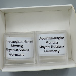 Könnte beinhalten: Zwei kleine, durchsichtige Plastikboxen mit weißen Etiketten. Die Etiketten haben schwarzen Text, der "ine-augite, richteri Mendig Mayen-Koblenz Germany" und "Aegirine-augite Mendig Mayen-Koblenz Germany" lautet.