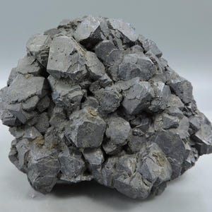 May include: A cluster of dark gray, metallic galena crystals featuring a cubic structure. The mineral specimen displays numerous interlocking, geometric shapes with sharp edges and a dull, lead-like luster across the entire surface of the rock.