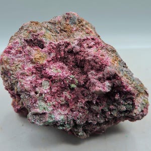 May include: A rough, irregularly shaped rock formation with a mix of colors. The dominant hues are deep pink and reddish-purple, with patches of green, brown, and gray. The surface has a textured, crystalline appearance.