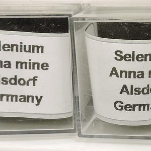 Op de afbeelding: Twee transparante, vierkante vitrines, elk met een wit etiket met zwarte tekst: "Selenium Anna mine Alsdorf Germany". De vitrines bevatten een donker materiaal, mogelijk een mineraal.