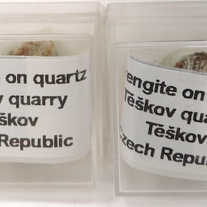 May include: Two clear plastic display cases each containing a small mineral sample. The samples are labeled "rengite on quartz, Těškov quarry, Těškov, Czech Republic."