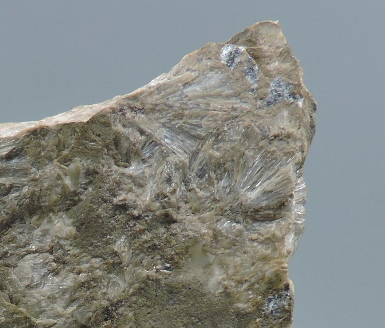 May include: Close-up of a rough, textured rock formation. The rock has a mix of gray and beige tones, with crystalline structures radiating outwards. The surface is uneven, with visible mineral deposits and a slightly weathered appearance.