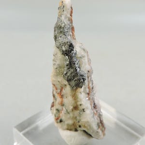 Può includere: Un campione di minerale con base chiara, inclusioni grigio scuro e marrone rossiccio. Il minerale ha una superficie ruvida e strutturata ed è montato su un supporto in acrilico trasparente. Formazione geologica naturale.