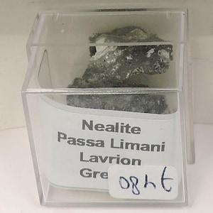 May include: A mineral specimen of Neaite from Passa Limani, Lavrion, Greece, encased in a clear plastic display box. The mineral is a dark gray color with white and green accents. A label with the mineral's name is visible.