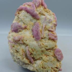 May include: A natural mineral specimen with a pale yellow and off-white stone matrix. It has several elongated, pinkish-purple crystal formations. The texture is coarse and uneven, highlighting the raw beauty of the geological formation. The specimen is a unique piece.
