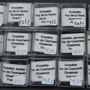 May include: A collection of 15 small, clear plastic boxes containing various mineral specimens. Each box has a label with the mineral name, location, and a number. The minerals include enstatite, pyroxene, biotite, amphibole, and hematite.