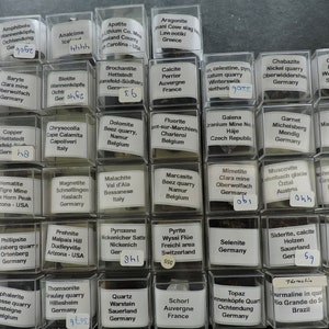 May include: A collection of 64 small, clear plastic boxes, each containing a different type of mineral specimen. The boxes are arranged in a grid of eight rows and eight columns. Each box has a white label with the name of the mineral, its location, and a number.