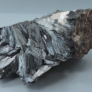 May include: A dark, metallic mineral specimen with a crystalline structure. The specimen has a dark gray, almost black color with a reflective, striated surface. The rough, brown matrix contrasts with the shiny, metallic crystals.