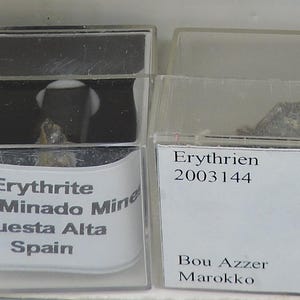 Op de afbeelding: Twee kleine, transparante plastic vitrines met mineraalmonsters. Eén kistje is gelabeld met "Erythrite, Cerro Minado Mine, Cuesta Alta, Spanje". De andere kist is gelabeld met "Erythrien 2003144, Bou Azzer, Marokko."
