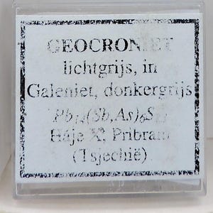 Op de afbeelding: Een kleine, doorzichtige plastic doos met een wit etiket met zwarte tekst. De tekst bevat het woord "GEOCRONIET" en andere wetenschappelijke termen, samen met de locatie "Pribram" en "Tsjechië".