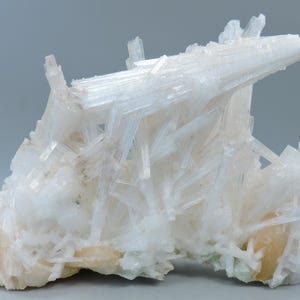 May include: A cluster of white, crystalline mineral formations. The crystals are elongated and needle-like, with a slightly translucent appearance. The base of the cluster has a pale, creamy color, with hints of orange and green.
