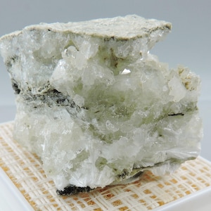 May include: A raw mineral specimen featuring a cluster of translucent, pale green and white crystalline structures. The rock displays a rough, jagged texture with visible internal layers of dark mineral inclusions throughout the crystalline mass.