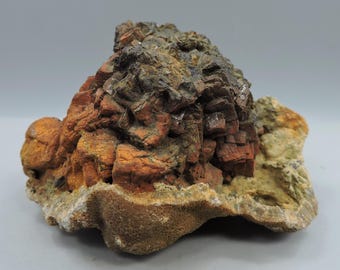 May include: A natural mineral specimen featuring a cluster of reddish-brown and dark brown crystalline structures. The mineral has a rough, textured surface with areas of orange and tan hues, and a base of light brown and beige.