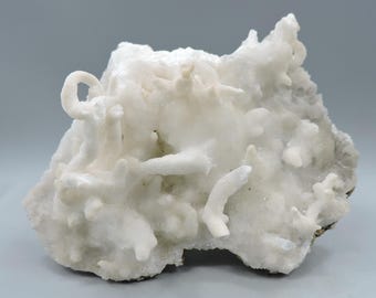 Op de afbeelding: Een wit, kristallijn mineraalspecimen met een ruw, getextureerd oppervlak. Het mineraal heeft een verscheidenheid aan vormen en maten, waaronder sommige die lang en dun zijn, en andere die ronder zijn. Het specimen is een natuurlijke formatie en heeft een unieke, organische uitstraling.