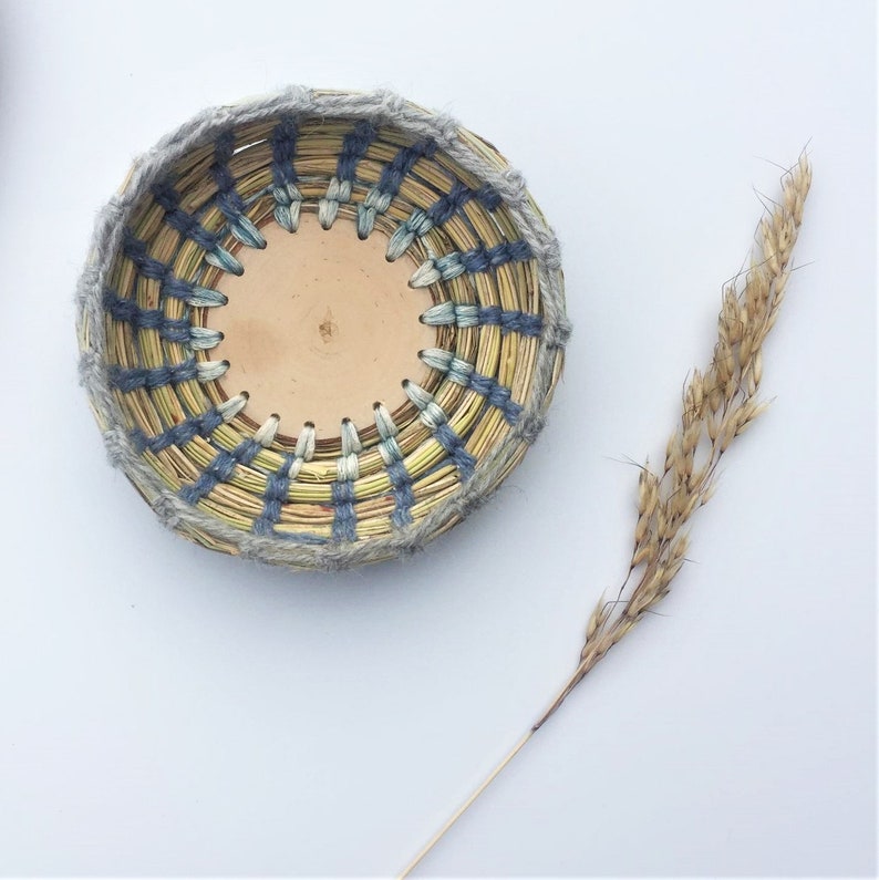 May include: A round woven basket with a light brown leather base. The basket is woven with natural fibres in shades of yellow, blue, and grey. The basket is sitting on a white surface.