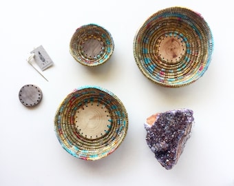 May include: Four woven baskets with colourful thread accents, a wooden disc, a tag, and a purple geode on a white background. The baskets have a natural wood base and are of varying sizes. The geode is a deep purple colour.