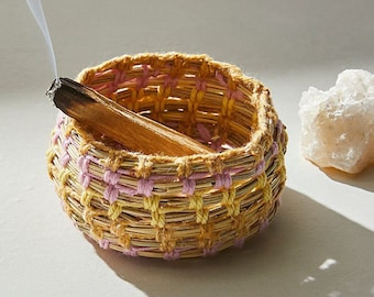 May include: A woven basket with yellow and pink stripes holds a smouldering stick of incense. The basket is made from natural materials and has a rustic, handmade look.