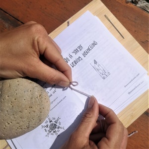 May include: A close-up of a person's hands tying a knot in a light pink string. The string is tied around a small stone. The person is following instructions from a book titled "Do it yourself weaving instructions".