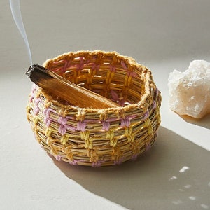 May include: A woven basket with pink and yellow accents holds a smoldering stick of incense. The basket is sitting on a white surface.