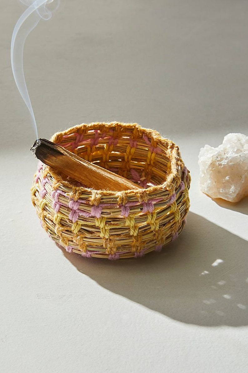 May include: A woven basket with pink and yellow accents holds a smoldering stick of incense. The basket is sitting on a white surface.