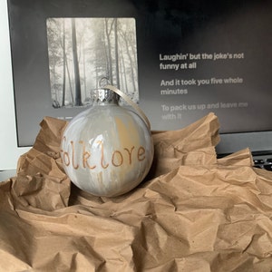 May include: A round, hand-painted Christmas ornament with the word "folklore" in copper-colored script. The ornament has a silver cap and a cream-colored ribbon. The background includes a laptop screen with song lyrics.