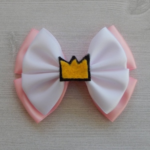 May include: A white and pink satin bow with a yellow felt crown in the center. The crown is outlined in black.