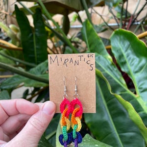 May include: Handmade crochet earrings in a rainbow color scheme. The earrings feature a chain link design with red, orange, yellow, green, blue, and purple yarn. The earrings are displayed on a brown tag with the text "Mirantics".