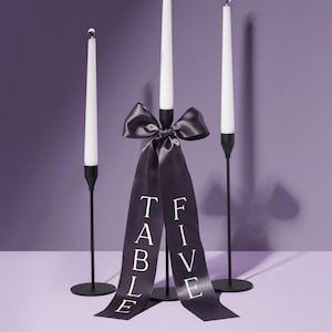 Könnte beinhalten: Drei schwarze Kerzenhalter mit weißen Kerzen. Ein schwarzes Satinband mit dem Text "TABLE FIVE" ist um den mittleren Kerzenhalter gebunden.