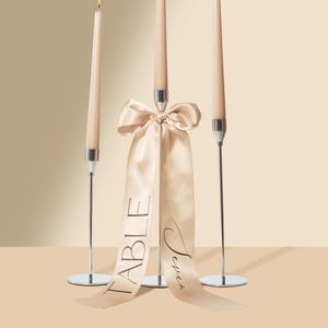 Könnte beinhalten: Drei brennende, schlanke, pfirsichfarbene Kerzen in silbernen Haltern. Ein pfirsichfarbenes Satinband mit den Worten "Table Seven" hängt von der mittleren Kerze. Auf neutralem Hintergrund.