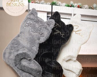 May include: Three plush cat-shaped Christmas stockings in gray, black, and white hang from a mantel. Each stocking has a name embroidered in gold thread. The background includes a fireplace with a fire.