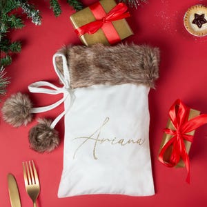 May include: A white Christmas stocking with a brown fur trim and the name "Ariana" in gold script. The stocking is decorated with two brown pom-poms and sits on a red surface with wrapped gifts and a fork and knife.