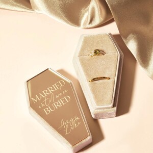 May include: A gold coffin-shaped ring box with the text "Married until we're buried" and the names "Amy & Luke" engraved on the lid. The box is open and shows a velvet interior with two rings.