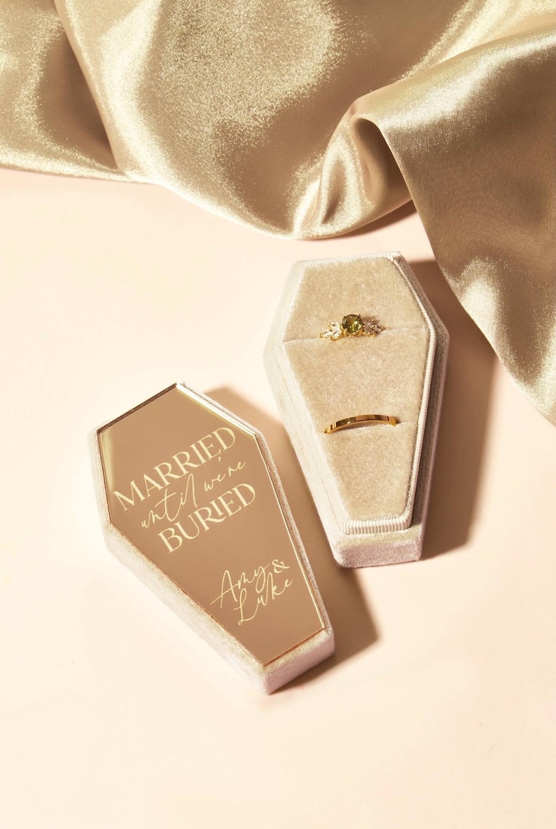 May include: A gold coffin-shaped ring box with the text "Married until we're buried" and the names "Amy & Luke" engraved on the lid. The box is open and shows a velvet interior with two rings.
