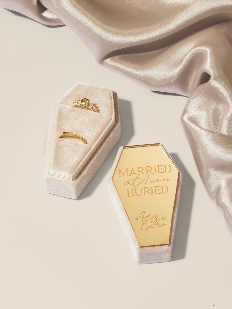 May include: Two gold rings in a white velvet coffin-shaped ring box. The box is engraved with the text "Married until we're buried" and the names "Amy & Luke".