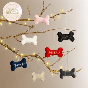 May include: A display of plush dog bone ornaments in various colors, including pink, white, red, navy, and gray, hanging from a gold-colored branch. Each bone has a name in gold script. The ornaments are for weddings and events.