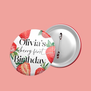 May include: A white button with a red border features a watercolor illustration of strawberries and the text "Olivia's berry first Birthday".